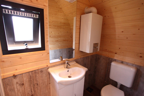 Baño con paredes y suelo de madera, lavabo, inodoro, espejo, calentador de agua y ventana en glamping.