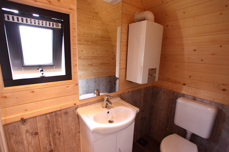 Baño con paredes y suelo de madera, lavabo, inodoro, espejo, calentador de agua y ventana en glamping.
