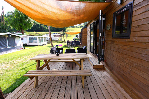 Terrazza in legno con tavolo da picnic davanti a una cabina glamping, ombreggiata e con roulotte sullo sfondo.