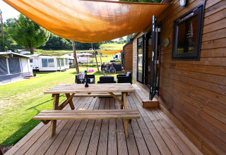 Træterrasse med bordbænk foran glampinghytte, skyggefuld overdækning og campingvogne i baggrunden.