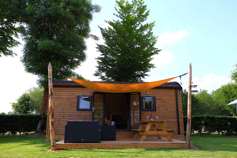 Cozy glamping cabin with wooden terrace, picnic table, and sunshade, set in a lush green campsite.