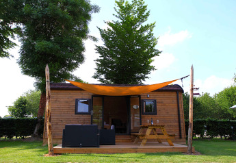 Gemütliches Glamping-Haus aus Holz mit Terrasse, Picknicktisch und Sonnensegel in grüner Umgebung.