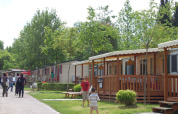 People walk and children play outside cozy wooden lodges surrounded by greenery and trees at a vacation park.