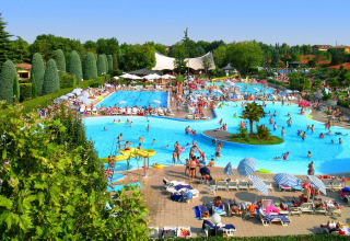 Gran área de piscina en una cabaña, llena de visitantes disfrutando bajo sombrillas en un día soleado.