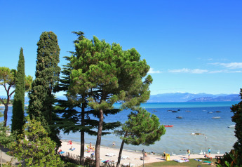 View from a lodge over a sunny beach, people swimming, green trees, and boats floating on a blue lake.