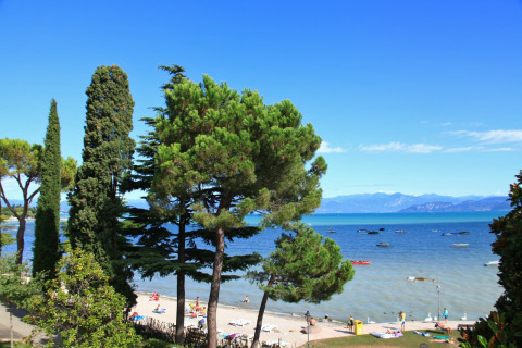 Vista da un lodge sulla spiaggia soleggiata, alberi verdi, persone e barche galleggianti sul lago azzurro.
