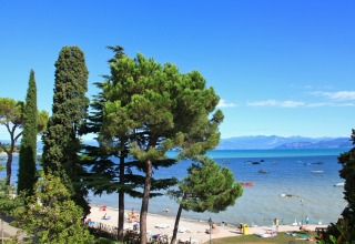 Vista da un lodge sulla spiaggia soleggiata, alberi verdi, persone e barche galleggianti sul lago azzurro.