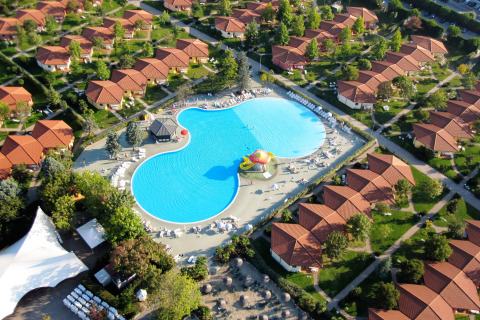 Luchtfoto van een vakantiedomein met rode dakbungalows en een groot centraal zwembad in het groen.