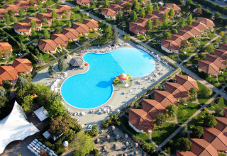 Luchtfoto van een vakantiedomein met rode dakbungalows en een groot centraal zwembad in het groen.