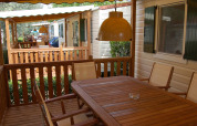 Houten terras bij een lodge met tafel, stoelen en hanglampen, omgeven door groene planten.