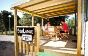 Dos personas desayunan en la terraza de madera de SunLodge, rodeados de árboles en un camping tranquilo.