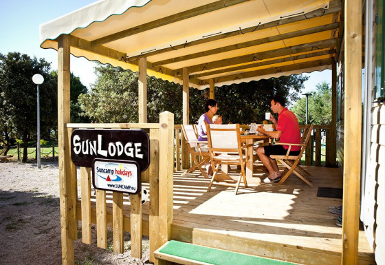 Dos personas desayunan en la terraza de madera de SunLodge, rodeados de árboles en un camping tranquilo.