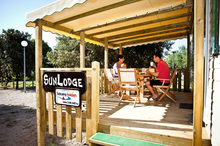 Dos personas desayunan en la terraza de madera de SunLodge, rodeados de árboles en un camping tranquilo.
