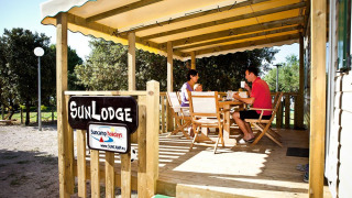 Two people have breakfast on the wooden SunLodge porch, surrounded by trees at a peaceful campsite.