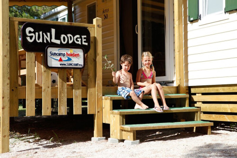 Twee kinderen zitten op de trap van een SunLodge met houten veranda onder een stralende zon.