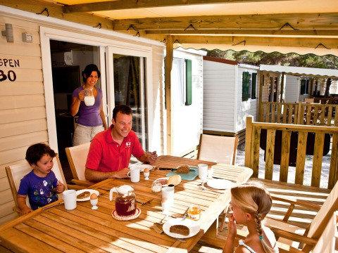 Gezin aan het ontbijt op houten terras aan een chalet in een vakantielodge, samen genietend buiten.