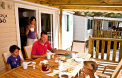 Family enjoys breakfast together on a wooden terrace outside a lodge cabin, smiling in the morning sun.