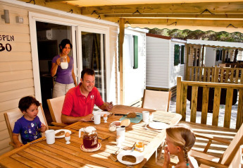 Familia desayuna junta en la terraza de madera de una cabaña, rodeada de otras casas en una lodge.