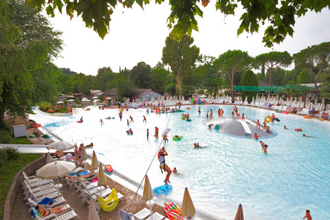 Piscina exterior en un lodge con niños y adultos jugando en el agua, rodeados de tumbonas y árboles verdes.