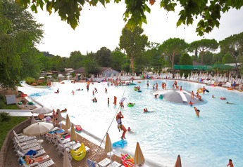 Outdoor swimming pool at a lodge with children and adults playing in the water, surrounded by sunbeds and trees.