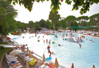 Udendørs swimmingpool ved en lodge, hvor børn og voksne leger i vandet omgivet af liggestole og træer.