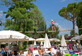 Child balancing on a rope adventure course above sun loungers and parasols at a lodge swimming pool.