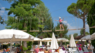 Bambino su percorso avventura sopra lettini e ombrelloni vicino alla piscina di un lodge all'aperto.