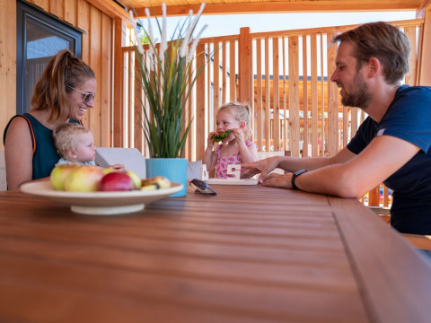 Una familia comparte una comida en una mesa de madera en la terraza de SunLodge Catalpa rodeada de madera.