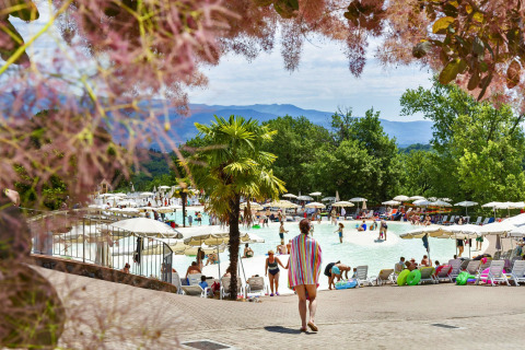 Person går mod en livlig udendørs swimmingpool omgivet af parasoller, grønne træer og bjerge i baggrunden.
