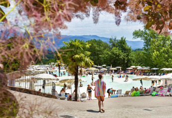 Person går mod en livlig udendørs swimmingpool omgivet af parasoller, grønne træer og bjerge i baggrunden.