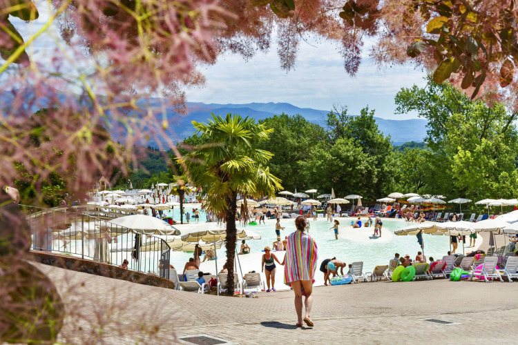 Une personne marche vers une piscine extérieure animée, entourée de parasols, d’arbres et de montagnes en fond.