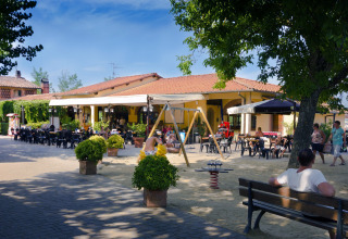 Terraza al aire libre en un lodge con personas sentadas, niños jugando y columpios bajo árboles frondosos.