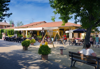 Udendørs terrasse ved en lodge med folk, borde, legende børn og legeredskaber under træer.