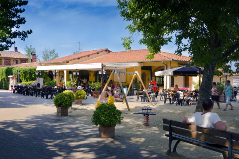 Udendørs terrasse ved en lodge med folk, borde, legende børn og legeredskaber under træer.