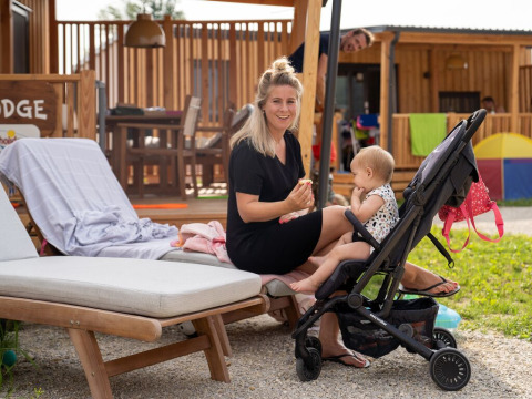 Vrouw met kindje in buggy geniet buiten bij SunLodge BigLeaf tegen houten lodge en terrasmeubilair.