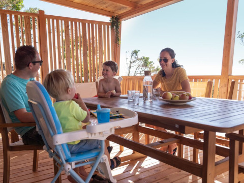 Une famille partage un repas sur la terrasse en bois ensoleillée du SunLodge BigLeaf, entourée de nature.