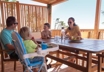 Een gezin geniet samen van een maaltijd op het zonnige houten terras van SunLodge BigLeaf, omringd door natuur.