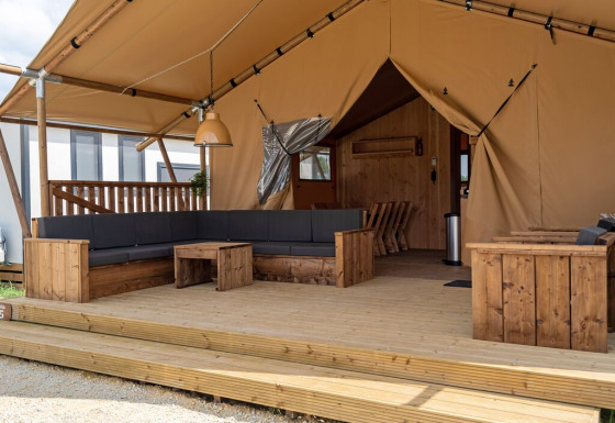 Ampia tenda glamping con veranda in legno, divano angolare e area relax in un campeggio moderno.