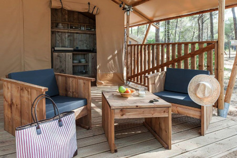 Zona de estar exterior con muebles de madera y cocina al fondo dentro de una tienda glamping en el bosque.