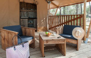 Zona de estar exterior con muebles de madera y cocina al fondo dentro de una tienda glamping en el bosque.