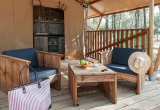 Zona de estar exterior con muebles de madera y cocina al fondo dentro de una tienda glamping en el bosque.