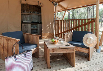 Zona de estar exterior con muebles de madera y cocina al fondo dentro de una tienda glamping en el bosque.