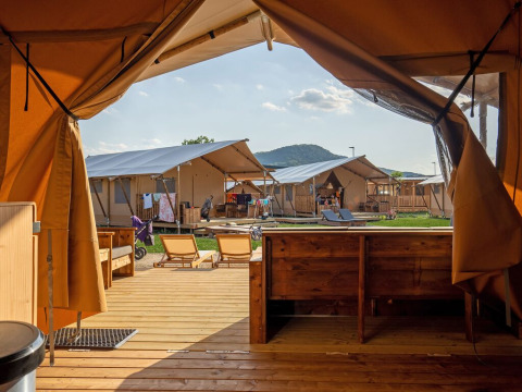 Uitzicht van binnenuit een luxe tent op een glamping met houten dek, ligstoelen en nabije tenten in de zon.