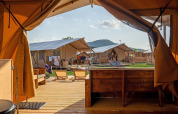 Uitzicht van binnenuit een luxe tent op een glamping met houten dek, ligstoelen en nabije tenten in de zon.