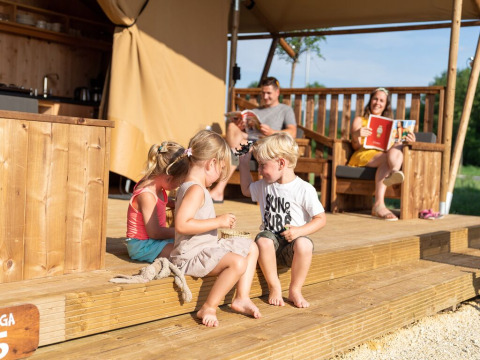 Niños juegan en la terraza de madera de SunLodge Taiga&Kambi glamping mientras los padres se relajan atrás.
