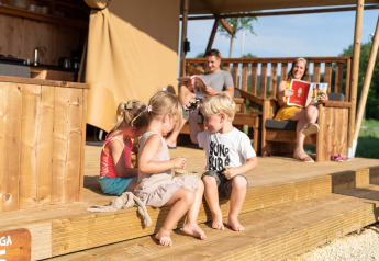 Kinderen spelen op het houten terras van SunLodge Taiga&Kambi glamping, ouders ontspannen op de achtergrond.