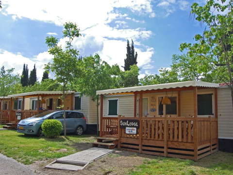 Foto van een SunLodge Maple lodge met veranda en wagen, omgeven door groen en een blauwe lucht.