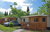 Foto van een SunLodge Maple lodge met veranda en wagen, omgeven door groen en een blauwe lucht.