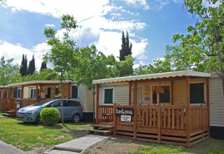 Foto van een SunLodge Maple lodge met veranda en wagen, omgeven door groen en een blauwe lucht.