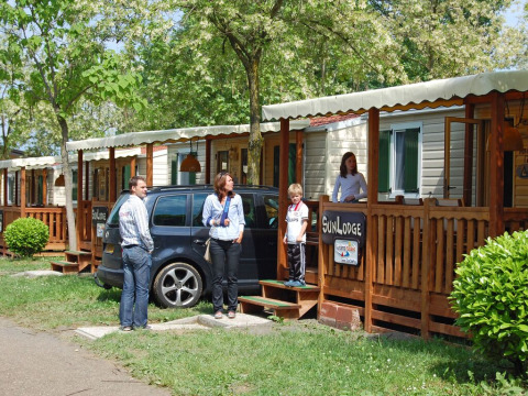 Famiglia davanti a SunLodge Maple a Camping Bella Italia in Italia, con auto e alberi vicino ai bungalow in legno.